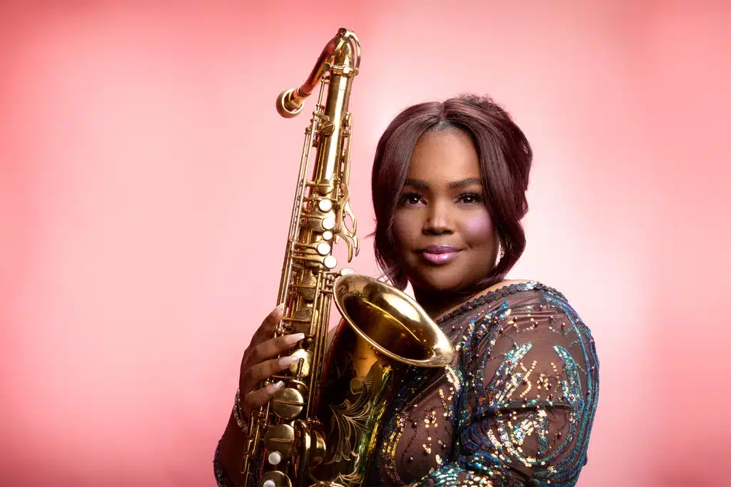 Camille Thurman holding a gold tenor saxophone in a sequined dress against a pink backdrop