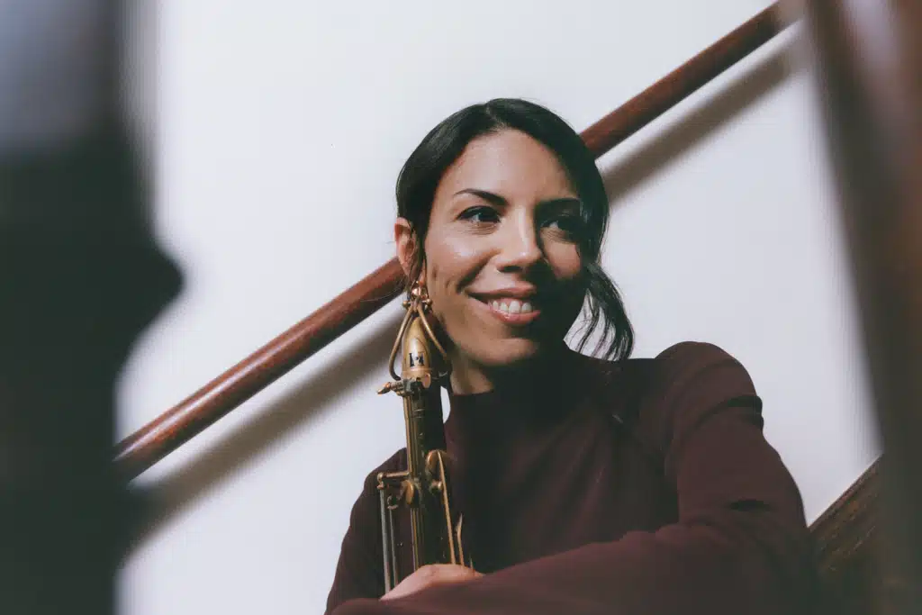 Melissa Aldana leaning on a stair rail holding her saxophone