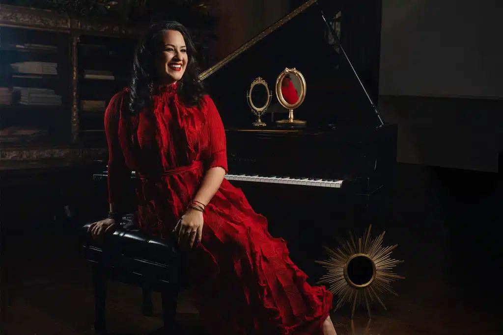 Nicole Zuraitis seated at a grand piano in a red dress, smiling warmly